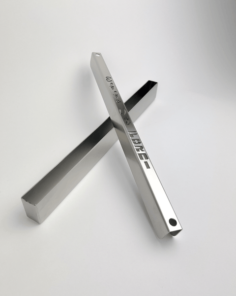 Zwei metallische Objekte liegen übereinander gekreuzt auf einem schlichten weißen Hintergrund. Ein Objekt, Teil des Effeuno Steamer - Dampferzeugers, ist ein glänzender, verspiegelter rechteckiger Balken mit kleinen Löchern und geprägtem Text, der an Dampferzeugerkomponenten erinnert. Das andere ist ein einfaches hohles rechteckiges Prisma mit polierter Oberfläche.