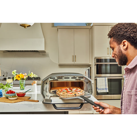 Eine Person nimmt in einer modernen Küche eine frisch gebackene Pizza aus einem Ooni Volt 12 Elektro Pizzaofen mit Pizzaschaufel. Die Arbeitsfläche vor ihnen ist mit einem Schneidebrett, einem Messer, einer Schüssel mit Kirschtomaten und einer Vase mit Blumen geschmückt.