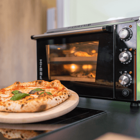 Im Vordergrund liegt auf einem hölzernen Pizzaschieber eine frisch gebackene Pizza mit Basilikumblättern. Im Hintergrund sieht man einen Effeuno Pizzaofen Effeuno P234H 509°C mit 2 Backkammern mit drei Drehreglern und einer transparenten Tür, in dem eine weitere Pizza gebacken wird, deren Rand perfekt auf einem Biscotto-Stein ruht.