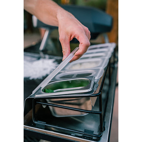 Eine Hand hebt den Deckel einer Ooni Pizza Topping Station von Ooni in einer Küche. Das Gerät enthält verschiedene Zutaten, darunter geschnittenes Gemüse, das teilweise sichtbar ist. Im Hintergrund sind weitere Kochgeräte zu sehen, die sich perfekt für die Zubereitung eines Pizza-Party-Buffets eignen.
