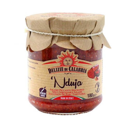 Un barattolo di "Delizie di Calabria Nduja Calabrese in barattolo (salame spalmabile)", un saporito paté spalmabile di maiale proveniente dalla Calabria, Italia. Il barattolo è chiuso con un coperchio di carta marrone legato con uno spago. L'etichetta indica che si tratta di un salame spalmabile da 180 g. Il disegno è in rosso e bianco con grafica al peperoncino.