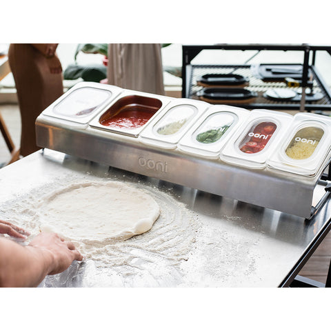 Eine Person formt einen Pizzateig auf einer bemehlten Fläche. Vor ihr steht eine Ooni Pizza Topping Station von Ooni mit durchsichtigen Deckeln mit der Aufschrift „Ooni“, die Zutaten wie Tomatensoße, Käse und verschiedene Beläge enthalten. Im leicht verschwommenen Hintergrund bereiten sie sich auf eine lustige Pizza-Party vor.