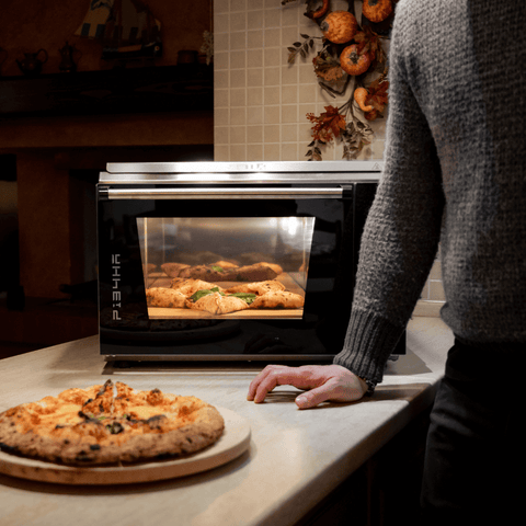 Eine Person steht in einer Küche neben einem mit Backwaren gefüllten Effeuno Pizzaofen Effeuno P134HA 509°C Evolution mit Biscotto Pizzastein. Im Vordergrund liegt eine frisch gebackene neapolitanische Pizza auf einem Holzbrett auf der Theke. Die Szene ist warm beleuchtet und im Hintergrund sind herbstliche Dekorationen zu sehen.