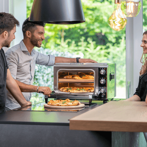 Drei Personen stehen um eine Arbeitsplatte herum, ein Effeuno Pizzaofen Effeuno P234H 509°C mit 2 Backkammern, in dem sich zwei Pizzen befinden. Ein Mann öffnet die Ofentür, während die anderen, ein Mann und eine Frau, lächeln und sich unterhalten. Die Küchenszene wird durch große Fenster im Hintergrund mit natürlichem Licht beleuchtet.