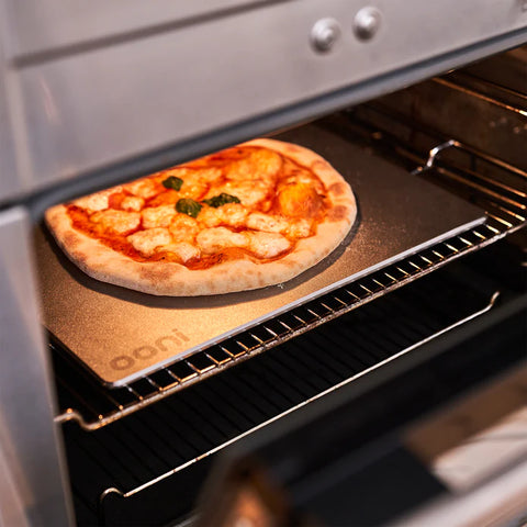 Eine mit geschmolzenem Käse und Basilikumblättern verzierte Pizza wird auf einem Ooni Pizzabackstahl 13 im Ofen gebacken. Die Ofentür ist teilweise geöffnet, so dass der Käse Blasen wirft und die knusprigen Pizzaböden anfangen, goldbraun zu werden.