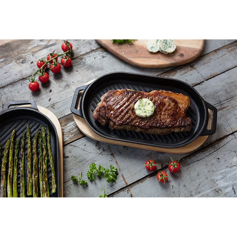 Ein gegrilltes Steak mit einer Scheibe Kräuterbutter liegt auf einer Ooni Grillpfanne aus Gusseisen und weist perfekte Grillstreifen auf. Daneben hält eine weitere Grillpfanne aus Gusseisen mit abnehmbarem Griff grüne Spargelstangen. Frische Kirschtomaten und Petersilie sind auf der rustikalen Holzoberfläche verstreut, zusammen mit einem Holzbrett, auf dem noch mehr Kräuterbutter liegt.