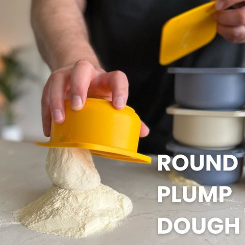 Eine Person hält ein Babadoh Silikon BIG Pizzateig und Zutatenbehälter 3er-Set, das auf einem Berg mit Mehl bestreuter Pizzateigkugeln ruht. Der Text „ROUND PLUMP DOUGH“ ist in klarer, weißer Schrift auf der rechten Seite des Bildes deutlich zu sehen. Im Hintergrund sind mehrere Schüsseln gestapelt, die die Szenerie für ein echtes Pizza-Abenteuer schaffen.