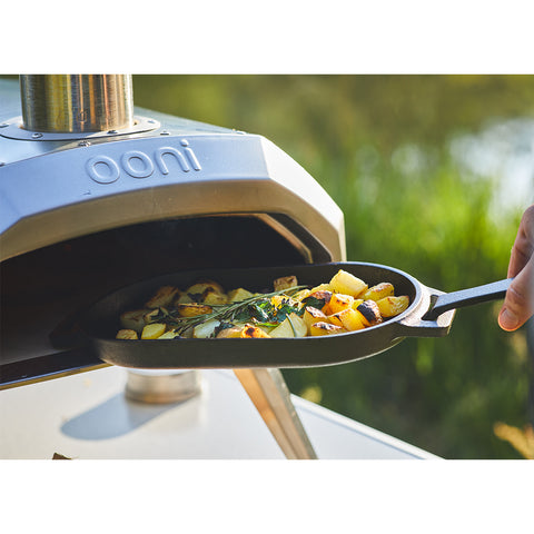 Eine Ooni Grillpfanne aus Gusseisen, gefüllt mit geröstetem Gemüse, darunter Kartoffeln und Blattgemüse, wird in einen Ooni Outdoor-Pizzaofen geschoben. Der abnehmbare Griff der Pfanne sorgt für mehr Komfort, während die Grillspuren den Geschmack verbessern. Der Ofen steht im Freien vor einem verschwommenen grünen Hintergrund.