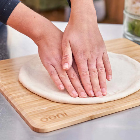 Zwei Hände drücken einen Pizzateig auf einen Ooni Teigdeckel & Vorbereitungsbrett aus nachhaltigem Bambus, der auf einem hölzernen Schneidebrett mit der Aufschrift „Ooni“ liegt. Im Hintergrund ist eine verschwommene Küchenkulisse zu sehen.