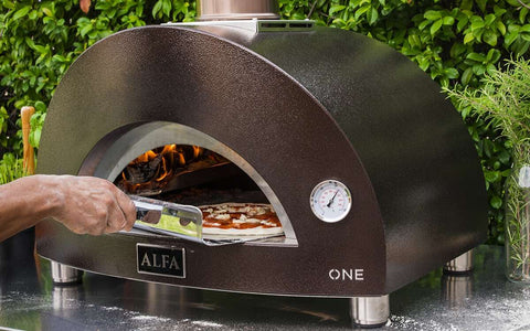 Eine Person schiebt eine neapolitanische Pizza auf einer Schaufel in einen eleganten, tragbaren Alfa Forni Moderno One Holz-Pizzaofen. Das schwarze Edelstahldesign verfügt über eine sichtbare Flamme und eine Temperaturanzeige auf der Vorderseite, im Hintergrund vor grünem Laub.