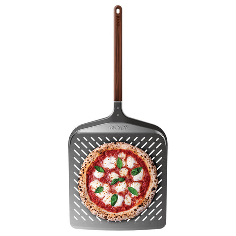 Eine frisch gebackene Pizza mit Mozzarella und Basilikum liegt auf dem Ooni Pizzaschieber Signature Perforiert 14" von Ooni, dessen Holzgriff von oben sichtbar ist.