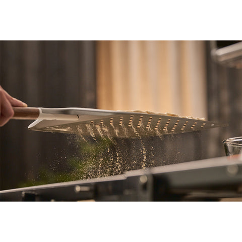 Eine Nahaufnahme des Ooni Pizzaschiebers Signature Perforiert 14" von Ooni, mit einem Holzgriff, der unter Teig und Mehlstaub in der Küche gleitet - perfekt für die Handhabung von Pizza wie ein Profi.