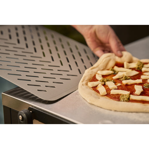 Eine Hand benutzt den Ooni Pizzaschieber Signature Perforiert 14" von Ooni, um eine rohe, belegte Pizza auf eine bemehlte Fläche zu schieben, bereit zum Backen.