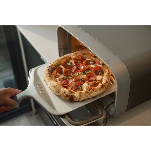 Eine Hand zieht mit dem Ooni Pizzaschaufel Kompakt 12" von Ooni mit innovativer FlourCatch-Technologie eine heiße Peperoni- und Jalapeno-Pizza aus einem kompakten Aufsatzofen.
