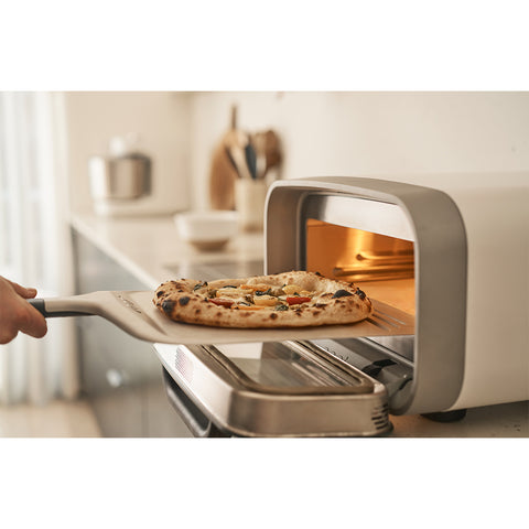 Eine Hand zieht eine frische Pizza aus einem modernen Arbeitsplattenofen mit der Ooni Pizzaschaufel Kompakt 12" von Ooni in einer hellen Küche, Küchenutensilien und Behälter verschwimmen sanft im Hintergrund.