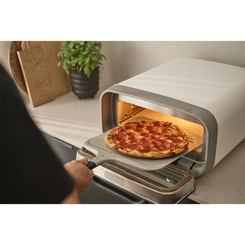 Mit der Ooni Pizzaschaufel Kompakt 12" von Ooni zieht eine Person eine Peperoni-Pizza aus einem Pizzaofen, während im Hintergrund eine Topfpflanze und Holzbretter zu sehen sind.