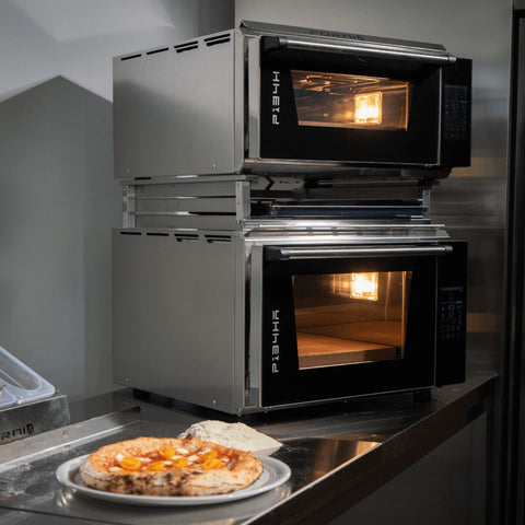 Zwei moderne Edelstahlöfen von Effeuno werden mit dem Effeuno Ofen Stapel-Kit auf einer Metalltheke gestapelt. Vorne zeigt ein Teller eine frisch gebackene Pizza, die mit orangenen Scheiben belegt ist, während beide Öfen mit Innenbeleuchtung leuchten.