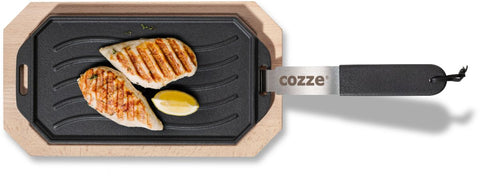 Gegrillte Hähnchenbrust und eine Zitronenspalte, serviert auf der Cozze Gusseisen-Wendepfanne mit Untersetzer 165x330mm, mit einem Metallgriff mit der Aufschrift "cozze" und einem Holzboden.