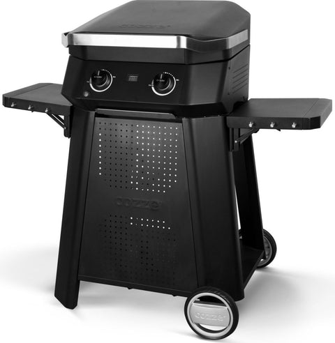 Ein schwarzer Cozze Gasgrill mit zwei Knöpfen, Metalldeckel, Seitenablagen, perforierter Front und Rädern passt zum Cozze Outdoor-Tisch für E-500 Elektrogrill, G-500 Gasgrill und Plancha. "Cozze"-Schriftzug auf Frontplatte und Rädern. Passt zu Pizzaofen Zubehör.