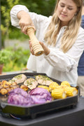 Eine Frau würzt Hähnchen, Mais, Kartoffeln und lila Kraut auf der Cozze G-500 Plancha mit Deckel (Gas 50mbar - CH-Regler) von Cozze. Der Grill steht im Freien, im Hintergrund ist Grün zu sehen.