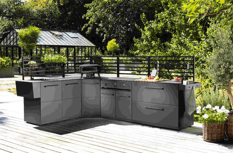 Eine moderne Outdoor-Küche mit schlichten schwarzen Schränken, Grill, Spüle und Küchenmodul 90 cm auf einer Holzterrasse im Grünen. Die Cozze Elements Rückwand für Outdoor-Küchenmodul [90 cm] von Cozze bietet Wind- und Wetterschutz.