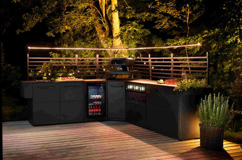 Eine Outdoor-Küche mit dunklen Schränken, eingebautem Grill, Mini-Kühlschrank und Platz für die Essenszubereitung auf einer Holzterrasse mit dem Cozze Elements LED-Rack für Outdoor-Küchenmodul (90 cm), beleuchtet von LED-Leuchten und umgeben von Bäumen.