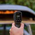 Eine Hand, die ein digitales Infrarot-Thermometer 2024 von Ooni hält, zeigt eine Temperatur von 500 Grad Fahrenheit an. Der Hintergrund ist leicht verschwommen und zeigt einen Pizzaofen im Freien mit einer sichtbaren Flamme im Inneren, perfekt zum Aufwärmen dieser Pizzasteine.