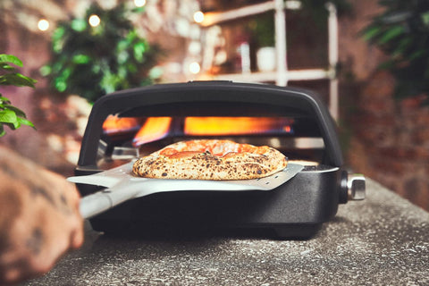 Eine Hand nimmt mit einem metallenen Pizzaschieber eine Pizza mit blubbernder Kruste aus dem Burnhard TONY Gas-Pizzaofen, dessen Flammen im Inneren flackern und hinter dem Grünzeug liegt, während die Pizza auf einem heißen Cordierit-Pizzastein ruht.