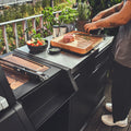 Eine Person bereitet auf einem Holzbrett neben dem Burnhard Beistellwagen (Black Edition) ein Steak und Kräuter zu, während Salat und frische Kräuter in einem stilvollen Kochbereich auf dem Balkon bereitstehen.