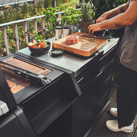 Eine Person bereitet auf einem Holzbrett neben dem Burnhard Beistellwagen (Black Edition) ein Steak und Kräuter zu, während Salat und frische Kräuter in einem stilvollen Kochbereich auf dem Balkon bereitstehen.