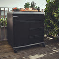 Der Burnhard Beistellwagen (Black Edition) von Burnhard steht auf einem Holzgestell, darauf ein Schneidebrett mit Brot und Gemüse, umgeben von Pflanzen – eine elegante Lösung für Ihren Outdoor-Küchenbereich.