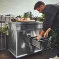 Ein Mann öffnet eine Schublade des „Burnhard Beistellwagen Edelstahl (Black Edition)“ von Burnhard, der auf einer Dachterrasse steht und mit Speisen und Kräutertöpfen beladen ist – umrahmt von üppigem Grün und Blick auf die Stadt, ideal für stilvolles Kochen im Freien.