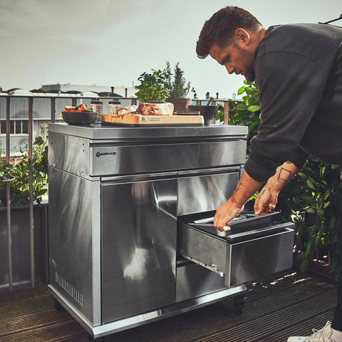 Ein Mann öffnet eine Schublade des „Burnhard Beistellwagen Edelstahl (Black Edition)“ von Burnhard, der auf einer Dachterrasse steht und mit Speisen und Kräutertöpfen beladen ist – umrahmt von üppigem Grün und Blick auf die Stadt, ideal für stilvolles Kochen im Freien.