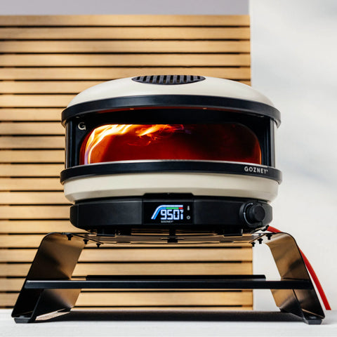 Ein Gozney ARC + ARC XL Booster steht in ergonomischer Höhe auf einem schwarzen Ständer und hat eine gut sichtbare digitale Temperaturanzeige mit „950 °F“. Der Ofen besticht durch ein modernes Design mit einer weißen und schwarzen Außenseite. Das Feuer im Inneren leuchtet hell und zeigt damit an, dass er in Betrieb ist – perfekt für jeden Pizza-Liebhaber.
