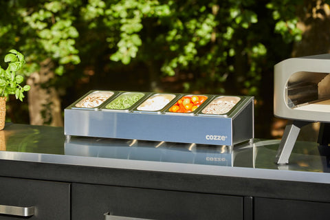 Die Cozze Pizza Topping Station Edelstahl von Cozze enthält geriebenen Käse, Mozzarellascheiben, Kirschtomaten und Champignons in vier GN-Behältern aus Stahl auf einer Arbeitsplatte im Freien neben einem Pizzaofen, vor der Kulisse üppiger grüner Bäume.