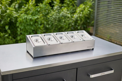 Die Cozze Pizza Topping Station Edelstahl von Cozze mit fünf abgedeckten Edelstahl-GN-Behältern wird auf einer modernen Außentheke präsentiert, umgeben von Grün und einem Metallzaun.