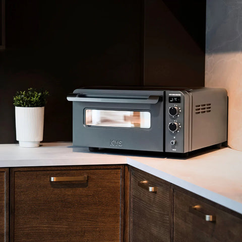 Der KYS Vero 500 Pro Elektro Pizzaofen steht auf einer Küchenarbeitsplatte neben einer weißen Topfpflanze, mit dunklen Holzschränken und einer hellen Aufkantung im Hintergrund.