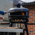Jemand schiebt eine Pizza in den Gozney Dome Pizzaofen (Multi-Fuel Gas/Holz) Off Black Bundle von Gozney, aus dem Flammen lodern. Der Ofen steht auf einem Sockel, unter dem Holz gelagert ist, vor einer Backsteinwand.