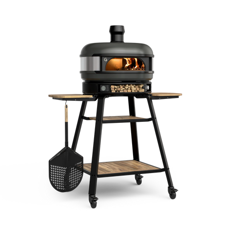 Ein Gozney Dome Pizzaofen (Multi-Fuel Gas/Holz) Off Black Bundle von Gozney wird auf einem Ständer mit Rädern und Holzregalen gezeigt, im Inneren brennt ein Feuer, an der Seite hängt ein Pizzaschieber und auf dem unteren Regal gelagertes Brennholz.