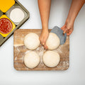 Ein Paar Hände bereitet gekonnt vier Teigbällchen auf einem bemehlten Holzbrett zu. Dazu verwendet es einen ergonomischen Babadoh-Teigschaber (Silikon). Daneben befindet sich ein Tablett mit Fächern, auf dem Tomatensoße, Mehl und andere Zutaten aufbewahrt werden.