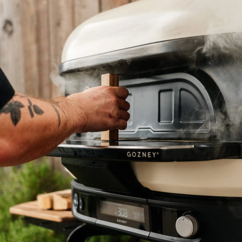 Eine Person mit einem Tattoo auf dem Arm öffnet die Gozney Pizzaofentür für Dome XL (Gen 2) von Gozney auf einem rauchenden Außenofen, umgeben von Holzstücken und Grünzeug.