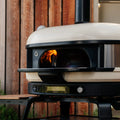 Ein Gozney Dome (Gen 2) Outdoor-Pizzaofen mit einem Feuer im Inneren steht auf einem schwarzen Metallständer mit der Gozney Ofensims & Frontablage vor einer Holzwand. Der Ofen verfügt über eine digitale Anzeige und das "Gozney"-Logo auf der Vorderseite.