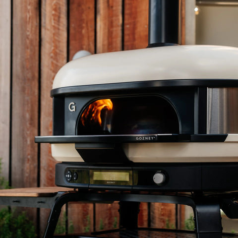 Ein Gozney Dome (Gen 2) Outdoor-Pizzaofen mit einem Feuer im Inneren steht auf einem schwarzen Metallständer mit der Gozney Ofensims & Frontablage vor einer Holzwand. Der Ofen verfügt über eine digitale Anzeige und das "Gozney"-Logo auf der Vorderseite.
