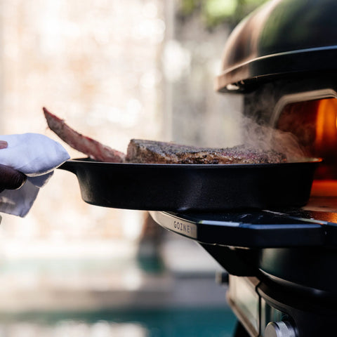 Mit einem Tuch wird eine heiße Pfanne mit einem großen, gewürzten Steak in den Gozney Ofensims & Frontablage für Dome XL (Gen 2) Outdoor-Ofen von Gozney gelegt, wo eine offene Flamme das Steak gart, während Dampf aufsteigt.