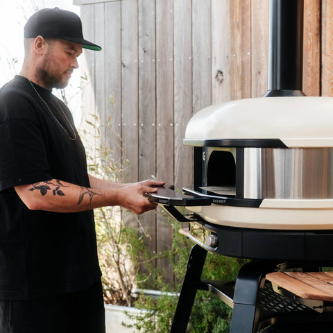 Ein Mann in schwarzem Hemd und Mütze macht sich bereit, einen Gozney Ofensims & Frontablage für Dome (Gen 2) Pizzaofen von Gozney auf einer modernen Terrasse mit Holzverkleidung, üppigen Pflanzen und dem praktischen Frontablage-Zubehör im Hintergrund zu öffnen.