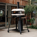 Ein Gozney Dome 2.0 Outdoor-Pizzaofen mit sichtbarer Flamme steht auf dem Gozney Stand & Pizzaofentisch für Dome 2.0 und Dome 2.0 XL, positioniert auf einer Terrasse neben einem Holzhaus und Topfpflanzen.