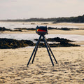 Ein Vermessungsgerät auf einem Stativ ist an einem Sandstrand mit Felsen und Wasser dahinter aufgestellt. Der bewölkte Himmel mit entfernten Bäumen und sanften Wellen umrahmt die Szene, ergänzt durch den Gozney Tread Gas-Pizzaofen Schwarz (mobil), der der Outdoor-Küche vor der ruhigen Naturkulisse Charme verleiht.