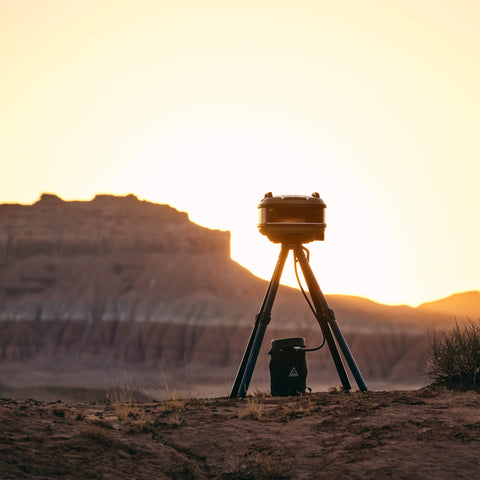 Ein auf einem Stativ montierter Gozney Tread Gas Pizzaofen Olive (Mobil) steht als Silhouette vor einem goldenen Sonnenuntergang in einer Wüstenlandschaft, perfekt für ein Outdoor-Küche-Erlebnis. Im Hintergrund ist eine Hochebene zu sehen, und trockene Vegetation bedeckt das Gelände, ideal für Camping- und kulinarische Enthusiasten.