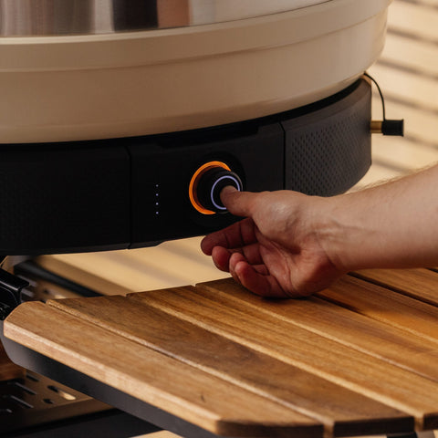 Eine Hand dreht den runden Luftstromsteuerungsknopf mit einer orangefarbenen LED auf dem Gozney Holzfeuer Control-Kit für Dome (Gen 2) / Dome XL (Gen 2), der über einer Holzlattenfläche angebracht ist.