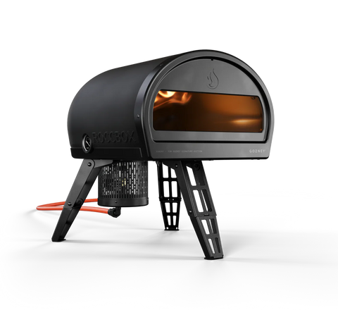 Un elegante e moderno forno a gas Gozney Roccbox di colore nero (Special Edition). Questo forno per pizza da esterno ha una forma cilindrica ed è sostenuto da gambe pieghevoli. L'apertura frontale rivela una fiamma visibile all'interno e promette una pizza di qualità da ristorante. Il logo e il marchio Gozney sono chiaramente visibili sul lato.